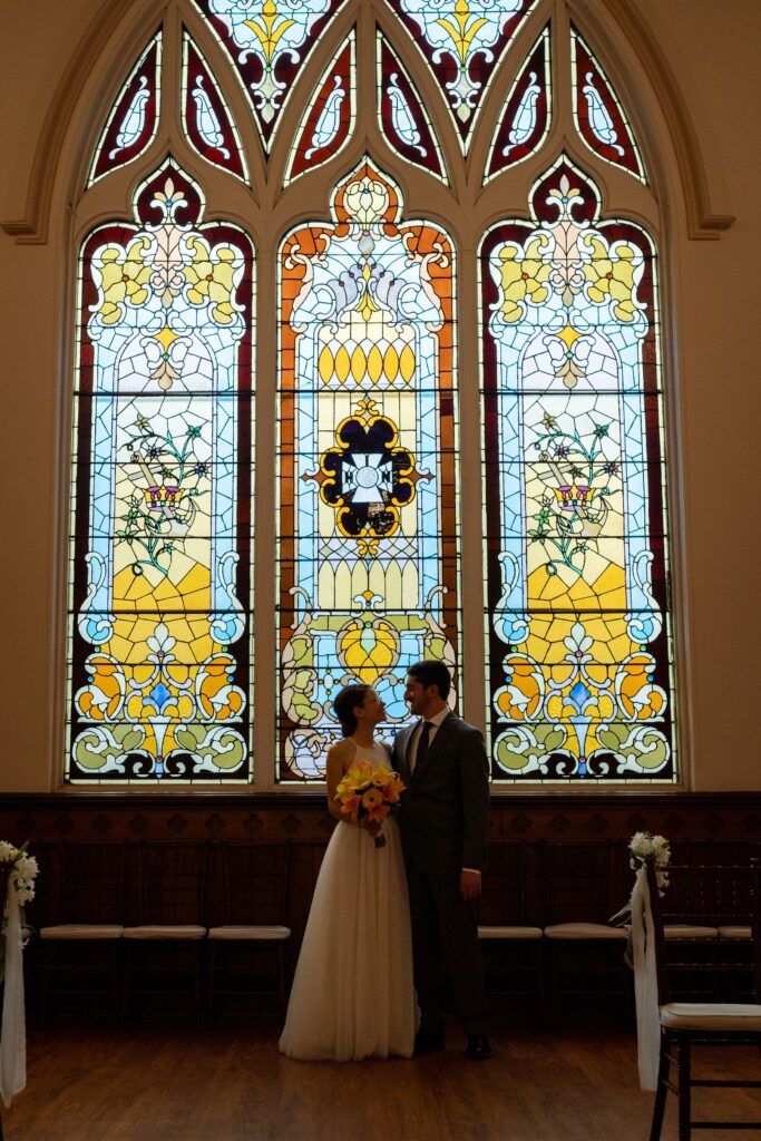A beautiful Rochester wedding. The bride is wearing a stunning white wedding gown with lace details, while the groom is dressed in a sharp black tuxedo. The wedding party, consisting of bridesmaids in blush pink dresses and groomsmen in black suits, are standing beside the couple. The ceremony is taking place in a charming church, with stained glass windows and wooden pews. The reception is held in a spacious venue decorated with elegant floral arrangements and romantic lighting. Guests are seen dancing, enjoying delicious food, and celebrating the love between the couple.