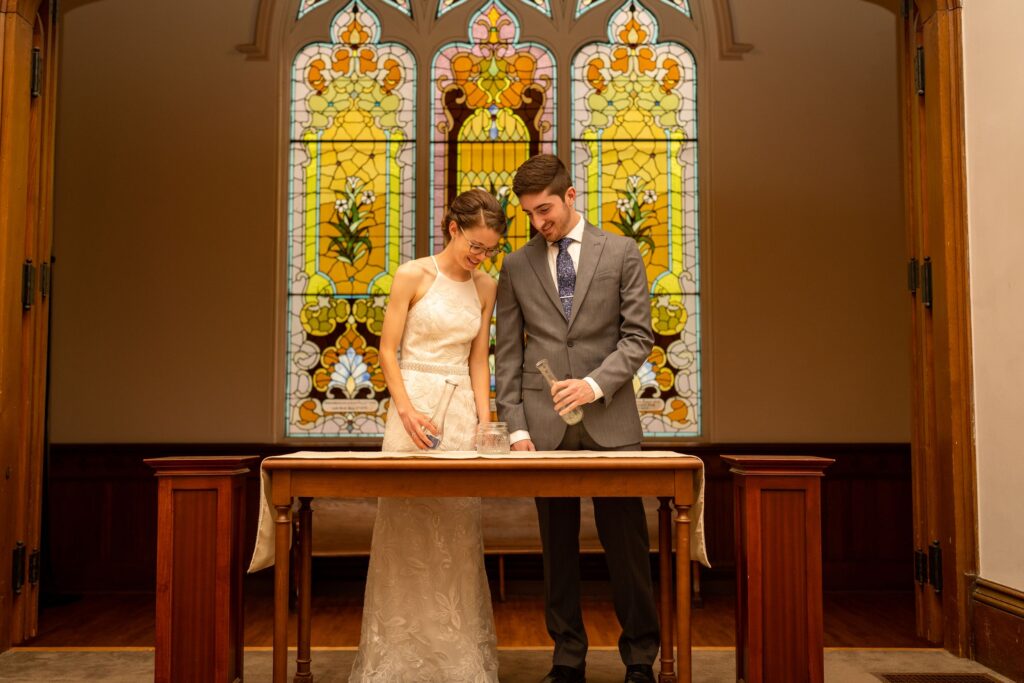 A beautiful Rochester wedding. The bride is wearing a stunning white wedding gown with lace details, while the groom is dressed in a sharp black tuxedo. The wedding party, consisting of bridesmaids in blush pink dresses and groomsmen in black suits, are standing beside the couple. The ceremony is taking place in a charming church, with stained glass windows and wooden pews. The reception is held in a spacious venue decorated with elegant floral arrangements and romantic lighting. Guests are seen dancing, enjoying delicious food, and celebrating the love between the couple.