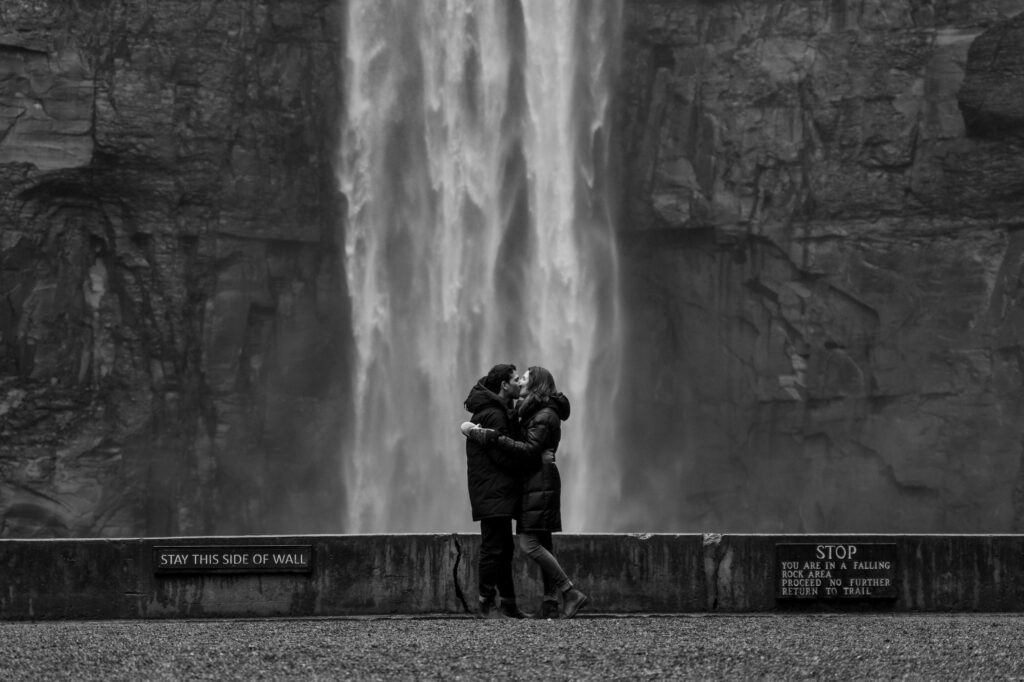 A man kneels down on one knee and holds out a ring box towards a woman with a stunning Taughannock waterfall as the backdrop. The proposal of Emma and Fady!