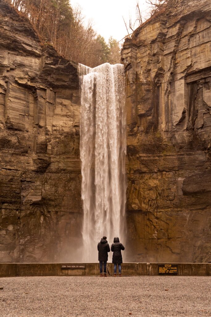 A man kneels down on one knee and holds out a ring box towards a woman with a stunning Taughannock waterfall as the backdrop. 