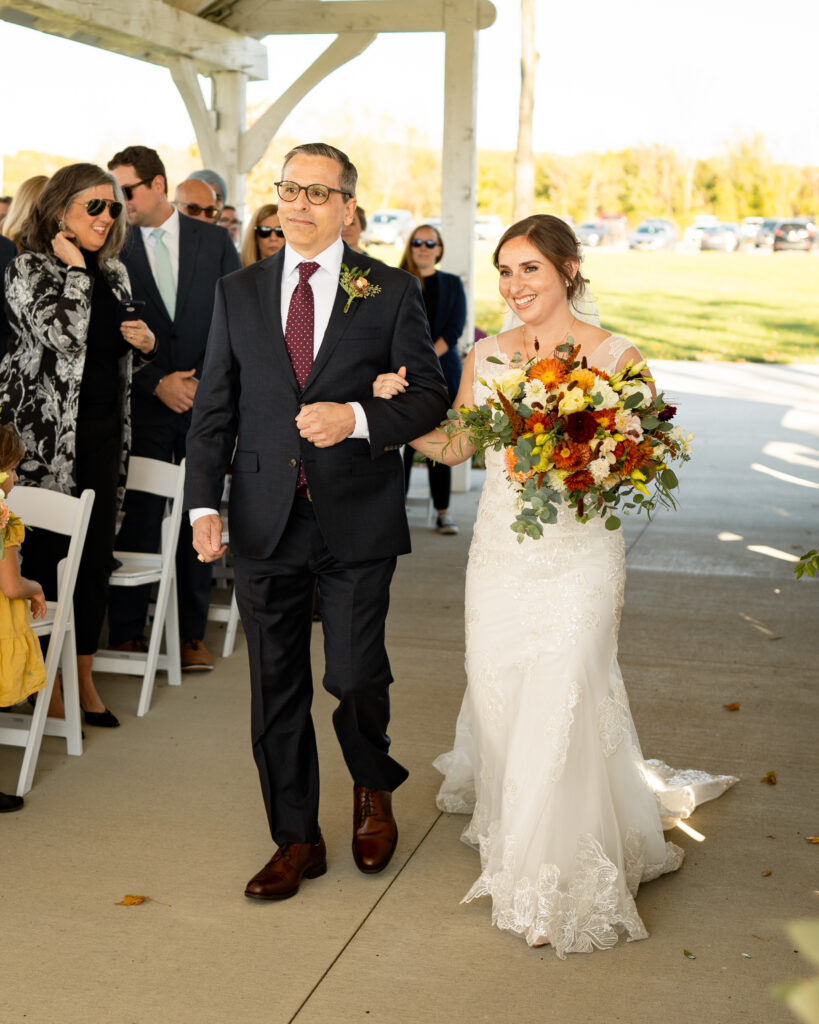 A scenic outdoor wedding ceremony in the fall. The bride, wearing a white wedding gown with lace details, is walking down an aisle covered with fallen leaves in shades of orange, yellow, and red. The groom, dressed in a dark suit, is standing under a beautiful arbor decorated with autumn foliage and flowers.