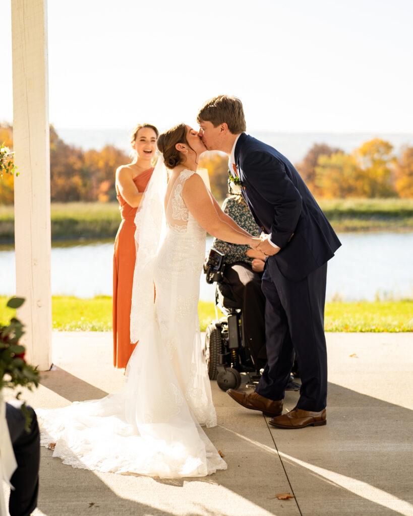 A scenic outdoor wedding ceremony in the fall. The bride, wearing a white wedding gown with lace details, is walking down an aisle covered with fallen leaves in shades of orange, yellow, and red. The groom, dressed in a dark suit, is standing under a beautiful arbor decorated with autumn foliage and flowers.