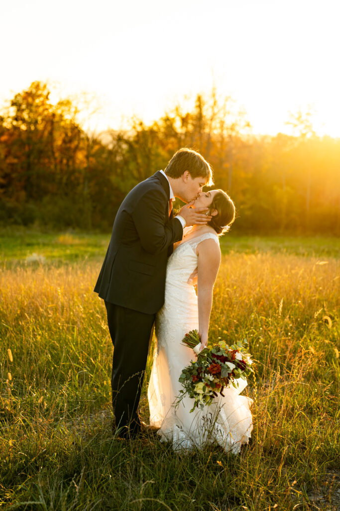 A romantic photo of the bride and groom walking hand in hand down a tree-lined path with leaves scattered on the ground. The bride's hair is styled in loose waves, and she's carrying a small bouquet of sunflowers and fall foliage.