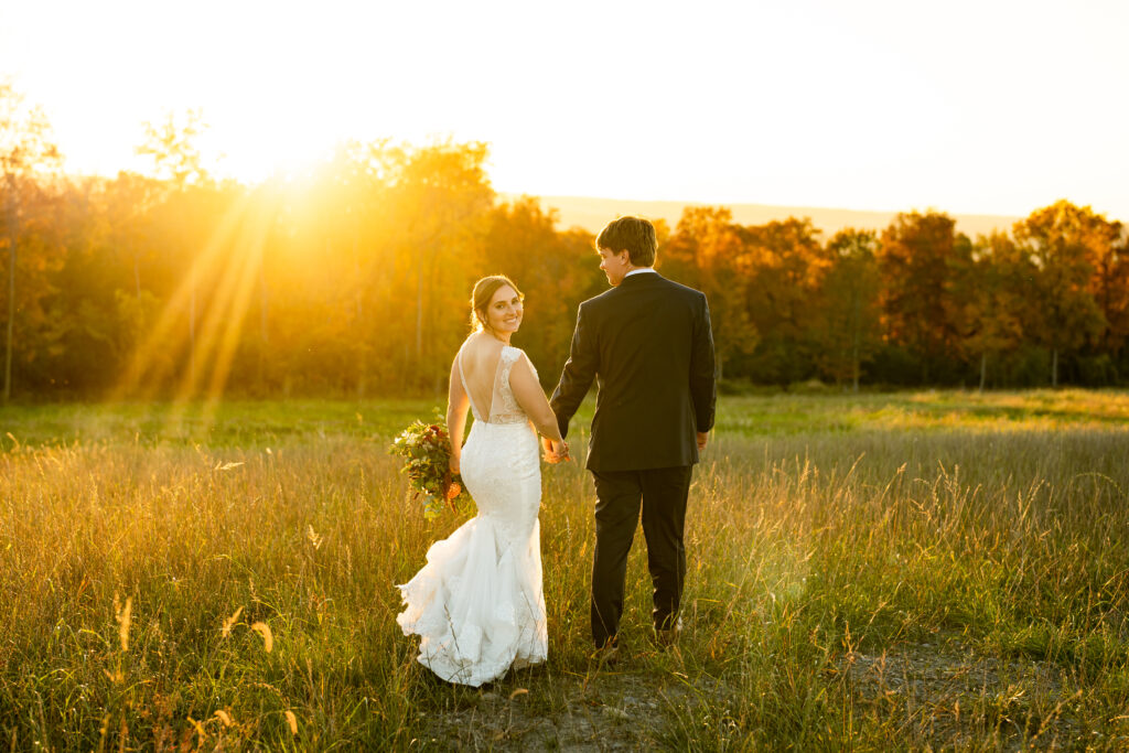 A romantic fall wedding photo of the bride and groom walking hand in hand down a tree-lined path with leaves scattered on the ground. The bride's hair is styled in loose waves, and she's carrying a small bouquet of sunflowers and fall foliage.