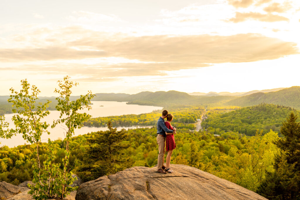 Engaged couple embraces in front of breathtaking mountain backdrop in the Adirondacks during their engagement photo session.