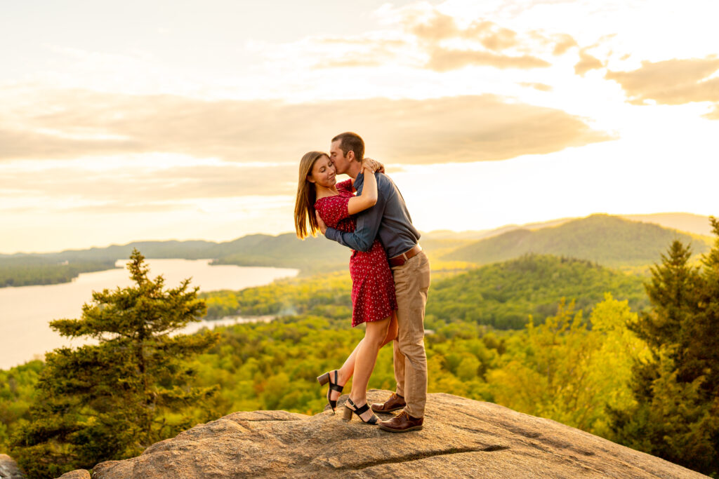 Engaged couple embraces in front of breathtaking mountain backdrop in the Adirondacks during their engagement photo session.
