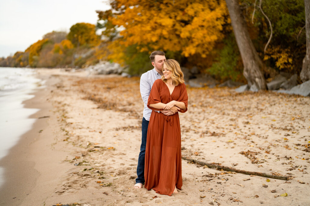 A beautiful fall beach engagement session at Durand Eastman Beach. The couple is walking hand in hand along the shoreline, with the colorful leaves of autumn providing a stunning backdrop. The bride-to-be is wearing a cozy sweater and jeans, while the groom-to-be is dressed in a casual shirt and pants. They are seen laughing and enjoying each other's company as they explore the beach and nearby woods. In some photos, they are sitting on a bench or standing on a wooden boardwalk, gazing into each other's eyes. The natural beauty of the fall landscape, combined with the couple's love and happiness, creates a magical atmosphere that is captured perfectly in the photographs.