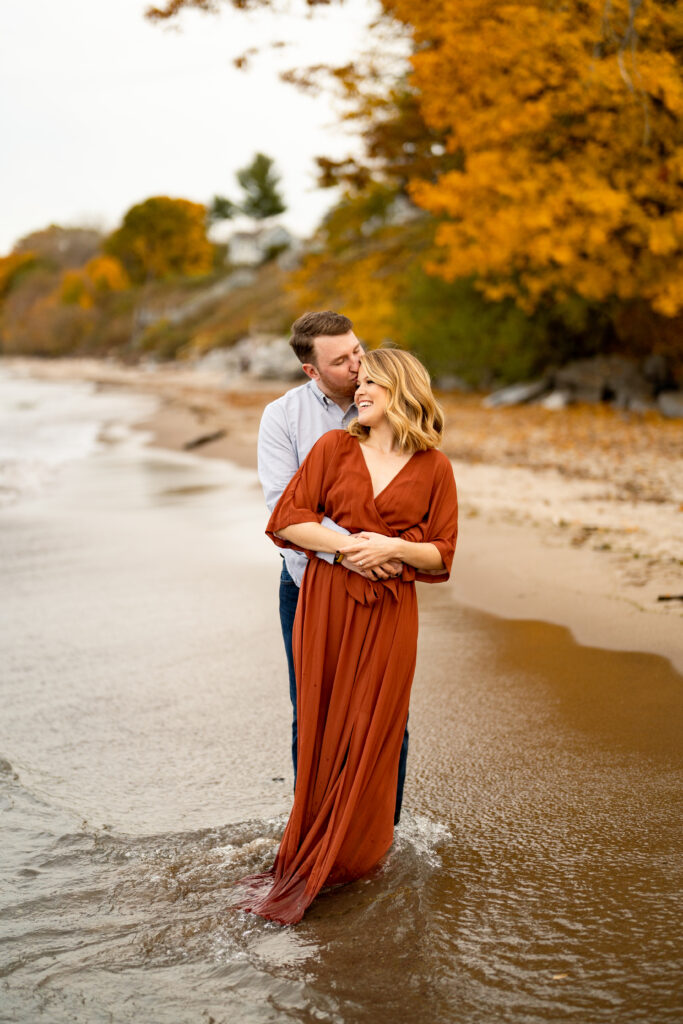 A beautiful fall engagement session at Durand Eastman Beach. The couple is walking hand in hand along the shoreline, with the colorful leaves of autumn providing a stunning backdrop. The bride-to-be is wearing a cozy sweater and jeans, while the groom-to-be is dressed in a casual shirt and pants. They are seen laughing and enjoying each other's company as they explore the beach and nearby woods. In some photos, they are sitting on a bench or standing on a wooden boardwalk, gazing into each other's eyes. The natural beauty of the fall landscape, combined with the couple's love and happiness, creates a magical atmosphere that is captured perfectly in the photographs.