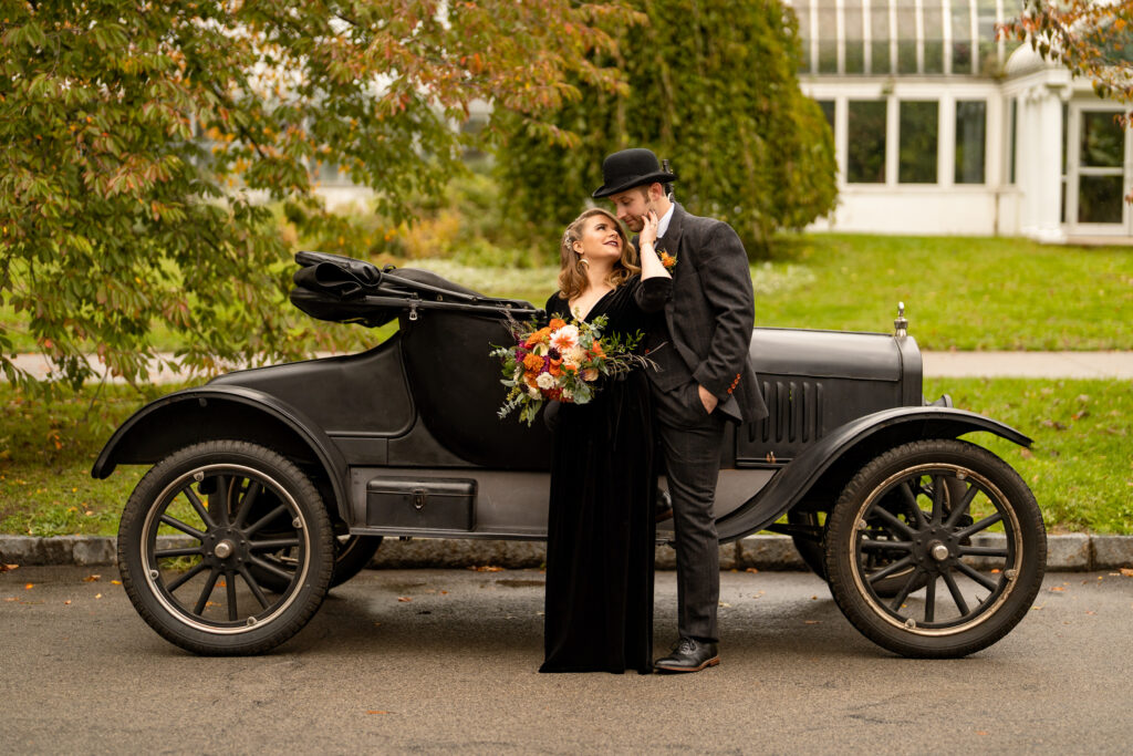 A Halloween wedding couple stands smiling in front of a vintage Model T car, posing for a photo to capture the essence of classic Americana. The bride, in a flowing black gown, stands delicately by the car door, while the groom, donning a dapper suit and tie, leans casually against the side of the vehicle. The antique car adds a touch of nostalgic charm to the photo, and the couple looks happy and excited to start their journey together, just as the Model T represents a symbol of new beginnings and adventure.