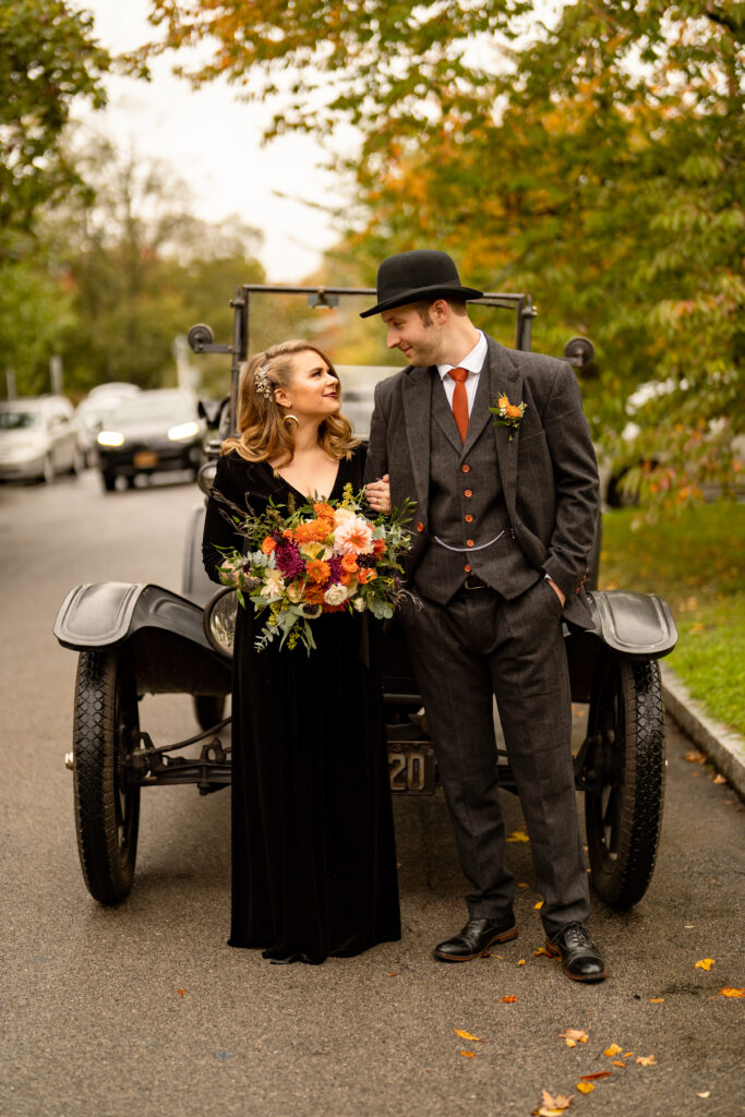 A Halloween wedding couple stands smiling in front of a vintage Model T car, posing for a photo to capture the essence of classic Americana. The bride, in a flowing black gown, stands delicately by the car door, while the groom, donning a dapper suit and tie, leans casually against the side of the vehicle. The antique car adds a touch of nostalgic charm to the photo, and the couple looks happy and excited to start their journey together, just as the Model T represents a symbol of new beginnings and adventure.