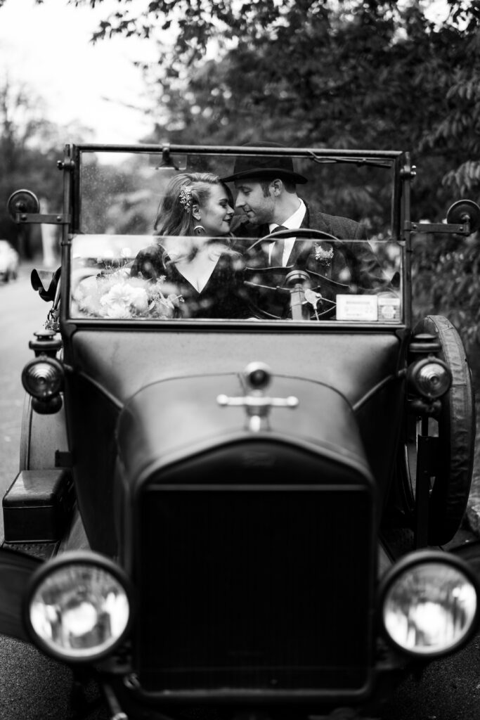 A wedding couple stands smiling in front of a vintage Model T car, posing for a photo to capture the essence of classic Americana. The bride, in a flowing black gown, stands delicately by the car door, while the groom, donning a dapper suit and tie, leans casually against the side of the vehicle. The antique car adds a touch of nostalgic charm to the photo, and the couple looks happy and excited to start their journey together, just as the Model T represents a symbol of new beginnings and adventure.