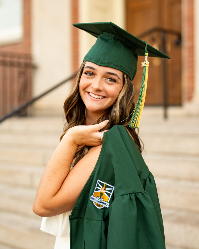 A college graduate proudly wears a cap and gown, posing for a photo session at SUNY Brockport. The graduate is standing in front of a brick building with trees in the background. The cap is square-shaped with a tassel hanging down the side, indicating the graduate's field of study. The gown is black with long sleeves and a V-shaped neckline, reaching down to the graduate's ankles. The graduate is smiling with a sense of accomplishment and holding a diploma scroll in one hand, while the other hand is raised in a celebratory gesture. The sun is shining, adding a warm and joyous atmosphere to the photo. The cap and gown represent the culmination of the graduate's hard work and dedication throughout their academic journey at SUNY Brockport.
