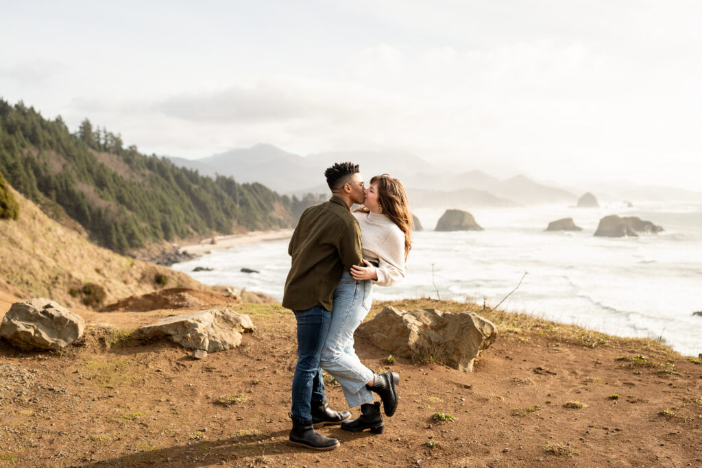 A couple stands close to each other, facing the ocean at Cannon Beach. The man has his arm around the woman's waist, and they are both looking out towards the horizon. The golden sunlight is casting a warm glow on their faces, and the waves crashing against the shore can be seen in the background. They are both smiling, lost in the moment of their intimate session on the beach, with the iconic Haystack Rock towering in the distance. Their love and connection are evident in their relaxed posture and happy expressions. 