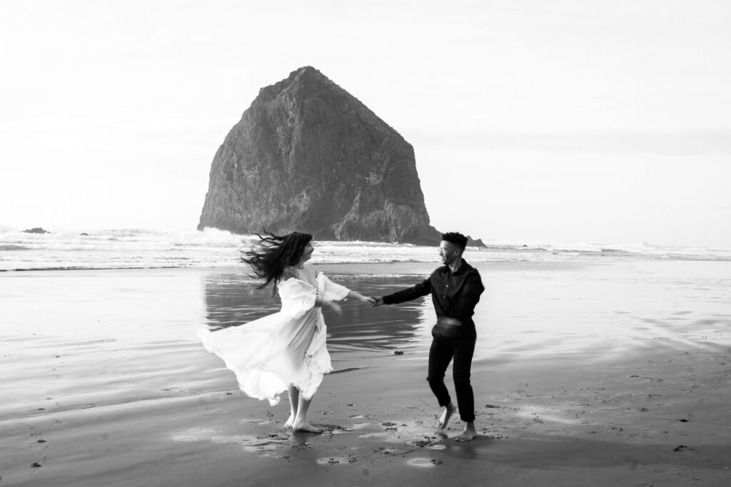 Nat and Luis walking hand in hand along the beach with Haystack Rock in the background, during their couples session at Cannon Beach.