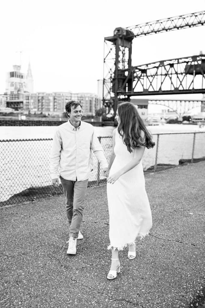 A happy couple, the woman in a white dress and the man in a light blue shirt and khaki pants, pose for romantic engagement photos in front of Lake Erie at the old coast guard station in Downtown Cleveland. The serene waters of Lake Erie provide a picturesque backdrop as they share a moment of love and connection, creating memories to last a lifetime.