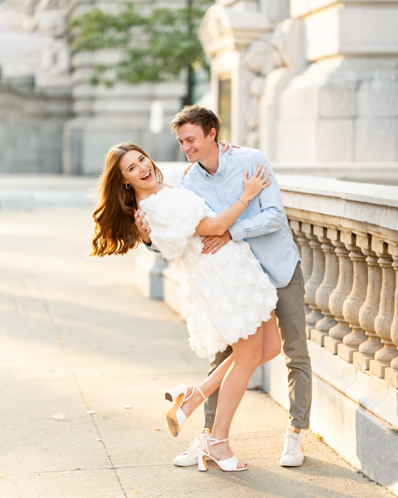 An affectionate couple, the woman dressed in a beautiful white gown and the man in a light blue shirt and khaki pants, share a tender moment while posing for engagement photos in front of the Downtown Cleveland Public Library. The love between them is palpable, and the iconic library building provides a stunning backdrop for their romantic journey.